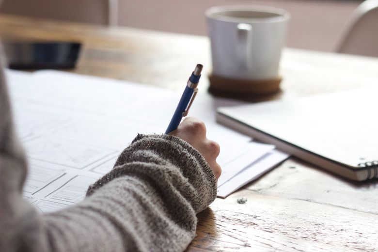 The arm of a student writing notes amid workbooks and a mug of coffee